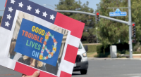 A person holds a sign at a street intersection with the phrase “GOOD TROUBLE LIVES ON” written in colorful letters next to a peace sign made of flowers. The sign is bordered in red, white, and blue with stars, resembling the American flag. In the background, a street sign reads “McBean Pkwy” and a white van waits at a red light.